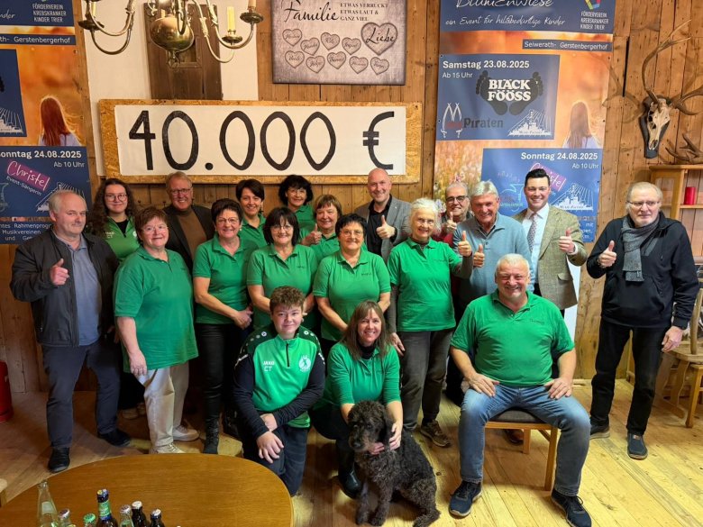 Spendenübergabe BLUMenwiese 2025 Eine größere Gruppe von Menschen steht in einem rustikal eingerichteten Raum zu einer Spendenübergabe zusammen. Viele tragen grüne Shirts und lächeln in die Kamera, einige zeigen Daumen nach oben. Hinter ihnen hängt ein großes Schild mit der Spendensumme „40.000 €“. Im Vordergrund sitzen und knien zwei Personen, eine davon mit einem Hund. Der Hintergrund ist dekoriert mit Veranstaltungsplakaten und Holzvertäfelung. Die Szene vermittelt eine feierliche und dankbare Atmosphäre anlässlich einer erfolgreichen Benefizveranstaltung.
