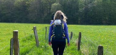 Wanderin mit Rucksack auf einem schmalen Wanderweg zwischen einer blühenden Wiese und einem Wald. Im Hintergrund sind ein bewölkter Himmel und eine weitläufige Frühlingslandschaft zu sehen.