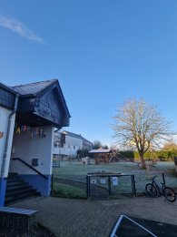Auf der linken Seite ist ein Kindergarterngebäude zu sehen mit dem Schriftzug "Rappelkiste". Rechts im Bild gibt es einen Spielplatz, der mit Spielgeräten ausgestattet ist, sowie eine große, kahle Baumkrone. Der Himmel ist strahlend blau, was einen schönen Kontrast zu den kalten Temperaturen und dem Frost auf dem Boden schafft. Diese Szenerie zeigt einen typischen Spielplatz in der Winterzeit.