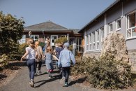 Das Bild zeigt eine Gruppe von Kindern, die fröhlich auf einem Weg in Richtung des KITA-Gebäudes gehen. Die Kinder sind bunt gekleidet und scheinen eine gute Zeit zu haben. Im Hintergrund ist eine helle, sonnige Umgebung mit viel grünem Pflanzenbewuchs zu sehen. Das Gebäude hat große Fenster und eine einladende Architektur. Die Atmosphäre wirkt freundlich und lebhaft, was auf eine positive Erlebnissituation hinweist.