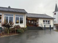 Das Bild zeigt das KITA-Gebäude von Außen. Ein modernes, einstöckiges Gebäude mit einem großen Eingangsbereich und mehreren Fenstern. Die Fassade ist in einem neutralen Farbton gehalten, und das Dach ist mit dunklen Schindeln gedeckt. Vor dem Gebäude gibt es wenige Pflanzen und eine Zugangstreppe. Im Hintergrund ist ein Kirchturm mit spitzem Dach sichtbar. Der Himmel wirkt grau und bewölkt.