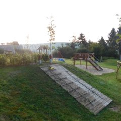 Das Bild zeigt einen Kinderspielplatz in einer ruhigen, grünen Umgebung. Im Vordergrund ist eine sanfte Hangfläche mit einer Holzrampe zu sehen. Auf der linken Seite befinden sich einige Sträucher oder kleine Bäume. Im Hintergrund gibt es mehrere Spielgeräte, darunter eine Rutsche und eine Kletterstruktur, sowie einen Sandkasten.