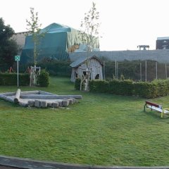 Das Bild zeigt einen weitläufigen Kinderspielplatz in einer grünen Umgebung. In der Mitte sind mehrere Spielgeräte angeordnet, darunter eine Kletterstruktur und Schaukeln. Es gibt zudem Sandkästen und eine Hüpfburg. Um den Spielbereich herum sind Bäume und Sträucher zu sehen, die für Schatten und eine naturnahe Atmosphäre sorgen.