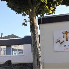 Das Bild zeigt die Fassade einer Kindertagesstätte, die hell und freundlich gestaltet ist. An der Wand ist ein bunter Schriftzug mit dem Namen „Kath. Kindertagesstätte Maria Himmelfahrt“ zu sehen. Rechts daneben sind spielerische Zeichnungen angebracht. Vor der Einrichtung steht ein großer Baum, der Schatten spendet. Der Himmel ist klar und blau, was eine einladende Atmosphäre vermittelt.