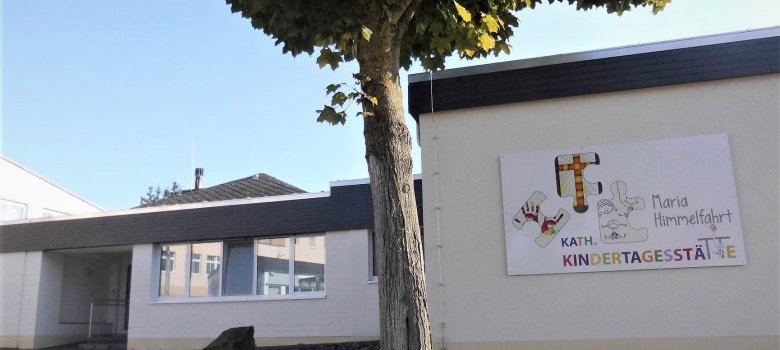 Das Bild zeigt die Fassade einer Kindertagesstätte, die hell und freundlich gestaltet ist. An der Wand ist ein bunter Schriftzug mit dem Namen „Kath. Kindertagesstätte Maria Himmelfahrt“ zu sehen. Rechts daneben sind spielerische Zeichnungen angebracht. Vor der Einrichtung steht ein großer Baum, der Schatten spendet. Der Himmel ist klar und blau, was eine einladende Atmosphäre vermittelt.
