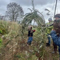 Ein Kind befindet sich in einer natürlichen Umgebung an einer Art Bau- oder Bastelprojekt beim Erstellen einer kleinen Struktur aus Zweigen oder Pflanzen. Mehrere Personen stehen dabei.