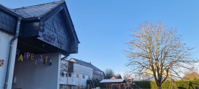 Auf der linken Seite ist ein Kindergarterngebäude zu sehen mit dem Schriftzug "Rappelkiste".  Rechts im Bild gibt es einen Spielplatz, der mit Spielgeräten ausgestattet ist, sowie eine große, kahle Baumkrone. Der Himmel ist strahlend blau, was einen schönen Kontrast zu den kalten Temperaturen und dem Frost auf dem Boden schafft. Diese Szenerie zeigt einen typischen Spielplatz in der Winterzeit.