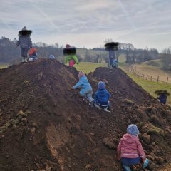Das Bild zeigt eine Gruppe von Kindern, die auf zwei großen Erdhügeln spielen und klettern. Die Kinder sind bunt gekleidet und scheinen Spaß zu haben, während sie sich an der frischen Luft in einer ländlichen Umgebung aufhalten. Die Landschaft im Hintergrund ist hügelig und grün, und der Himmel ist teilweise bewölkt. 