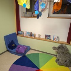 Das Bild zeigt ein gemütliches Spielzimmer. In der Ecke steht ein blauer Sitzsack mit einem lila Kissen. An der Wand sind verschiedene Spielzeug-Boxen und Bücher auf einem Regal angeordnet. Der farbenfrohe, runde Teppich mit einem Regenbogenmuster ergänzt die lebendige Stimmung des Raumes. In einer anderen Ecke steht ein grauer Plüschelefant.
