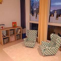 Das Bild zeigt einen hellen und einladenden Raum mit großen Fenstern, durch die sanftes Licht strömt. In der Ecke stehen zwei gepolsterte Sessel mit grüner Polsterung und weißen Punkten, die kindgerecht gestaltet sind. Neben den Sesseln befindet sich ein Regal mit mehreren Fächern, das mit Büchern und Spielen gefüllt ist. Die Fenster sind mit langen Vorhängen in Gelb und Blau dekoriert, was eine warme Atmosphäre schafft. Auf dem Boden liegt ein heller Teppich, der den Raum gemütlich erscheinen lässt.