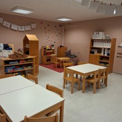 Das Bild zeigt einen freundlichen und hellen Raum in der KiTa Fleringen. Im Vordergrund stehen mehrere helle Tische mit Stühlen. An der Wand hängen bunte Zeichnungen und Dekorationen. Rechts befinden sich Regale mit verschiedenen Spielmaterialien, während in der Ecke ein kleiner Spielbereich mit weiteren Spielzeugen eingerichtet ist. Die Wände sind in einem sanften Rosa gestrichen, was dem Raum eine warme und einladende Atmosphäre verleiht.
