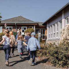 Das Bild zeigt eine Gruppe von Kindern, die fröhlich auf einem Weg in Richtung des KITA-Gebäudes gehen. Die Kinder sind bunt gekleidet und scheinen eine gute Zeit zu haben. Im Hintergrund ist eine helle, sonnige Umgebung mit viel grünem Pflanzenbewuchs zu sehen. Das Gebäude hat große Fenster und eine einladende Architektur. Die Atmosphäre wirkt freundlich und lebhaft, was auf eine positive Erlebnissituation hinweist.