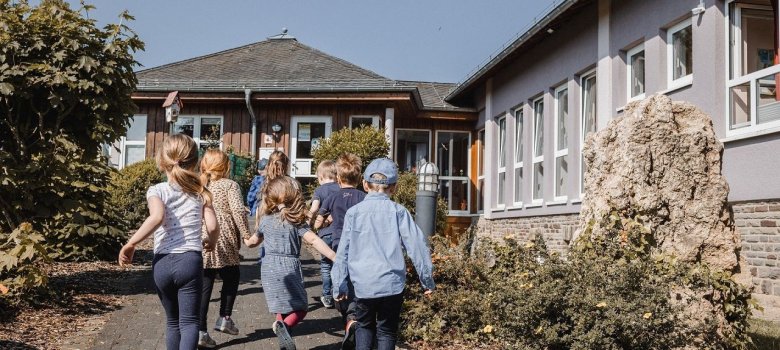 Das Bild zeigt eine Gruppe von Kindern, die fröhlich auf einem Weg in Richtung des KITA-Gebäudes gehen. Die Kinder sind bunt gekleidet und scheinen eine gute Zeit zu haben. Im Hintergrund ist eine helle, sonnige Umgebung mit viel grünem Pflanzenbewuchs zu sehen. Das Gebäude hat große Fenster und eine einladende Architektur. Die Atmosphäre wirkt freundlich und lebhaft, was auf eine positive Erlebnissituation hinweist.