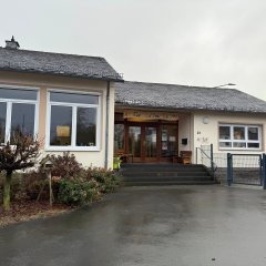 Das Bild zeigt das KITA-Gebäude von Außen. Ein modernes, einstöckiges Gebäude mit einem großen Eingangsbereich und mehreren Fenstern. Die Fassade ist in einem neutralen Farbton gehalten, und das Dach ist mit dunklen Schindeln gedeckt. Vor dem Gebäude gibt es wenige Pflanzen und eine Zugangstreppe. Im Hintergrund ist ein Kirchturm mit spitzem Dach  sichtbar. Der Himmel wirkt grau und bewölkt.