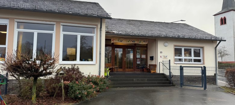 Das Bild zeigt das KITA-Gebäude von Außen. Ein modernes, einstöckiges Gebäude mit einem großen Eingangsbereich und mehreren Fenstern. Die Fassade ist in einem neutralen Farbton gehalten, und das Dach ist mit dunklen Schindeln gedeckt. Vor dem Gebäude gibt es wenige Pflanzen und eine Zugangstreppe. Im Hintergrund ist ein Kirchturm mit spitzem Dach  sichtbar. Der Himmel wirkt grau und bewölkt.