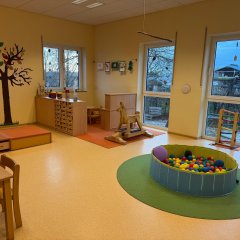Das Bild zeigt einen hellen Kindergartenraum mit gelben Wänden. Es gibt Tische, Stühle, ein Schaukelpferd und einen bunten Ballpool auf einem runden Teppich. An der Wand ist ein großer Baum gemalt, und die Fenster lassen viel Licht herein.