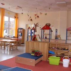 Der Raum ist hell gestaltet. Es gibt mehrere Holztische und Stühle, die um die Wände angeordnet sind. An den Wänden hängen dekorative Elemente wie beispielsweise Schmetterlinge oder andere Bastelarbeiten. In der Mitte des Raumes steht eine große Spielzeugkiste aus Holz, die verschiedene Spielzeuge enthält, darunter Fahrzeuge. Die Fenster lassen viel natürliches Licht herein, das den freundlichen und einladenden Charakter des Raumes verstärkt. Zudem gibt es einen Teppichbereich mit weichen Matten zum Spielen und Entspannen.