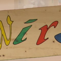 Das Bild zeigt ein handgemaltes Schild mit dem Schriftzug „Kita Wirbelwind“. Die Buchstaben sind in verschiedenen, lebendigen Farben gehalten, darunter Rot, Blau, Gelb und Grün, und wirken fröhlich und einladend. Das Schild hat eine geschwungene Form und ist aus Holz gefertigt, was einen kreativen und kindgerechten Eindruck vermittelt.