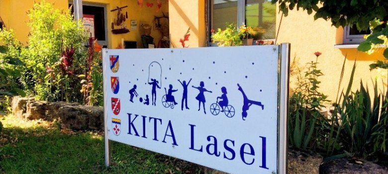 Das Bild zeigt ein Schild mit der Aufschrift „KITA Lasel“, das vor einem hellen Gebäude aufgestellt ist. Umgeben von grünem Gras und blühenden Pflanzen, vermittelt das Schild einen einladenden Eindruck. Nach oben hin sind Silhouetten von spielenden Kindern zu sehen, die Aktivitäten wie Radfahren und Tanzen darstellen. Das Gesamtbild strahlt eine fröhliche und lebendige Atmosphäre aus, passend für eine Kindertagesstätte.