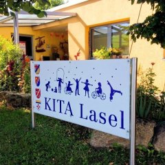 Schild mit der Aufschrift „KITA Lasel" Das Bild zeigt ein Schild mit der Aufschrift „KITA Lasel“, das vor einem hellen Gebäude aufgestellt ist. Umgeben von grünem Gras und blühenden Pflanzen, vermittelt das Schild einen einladenden Eindruck. Nach oben hin sind Silhouetten von spielenden Kindern zu sehen, die Aktivitäten wie Radfahren und Tanzen darstellen. Das Gesamtbild strahlt eine fröhliche und lebendige Atmosphäre aus, passend für eine Kindertagesstätte.