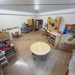 Das Bild zeigt einen weitläufigen Raum, der als Spiel- und Lernbereich für Kinder dient. In der Mitte steht ein runder Tisch auf Rädern, der umgeben ist von verschiedenen Regalen und Aufbewahrungseinheiten aus Holz, die sorgfältig organisiert sind. An den Wänden sind Zeichnungen und bilderhängende Elemente sichtbar. Eine Ecke des Raumes enthält einen Kletterbereich mit bunten Elementen, während ein weiterer Bereich eine gemütliche Sitzecke mit Kissen bietet. Die helle Raumgestaltung und die Vielzahl an Spielmaterialien schaffen eine freundliche und anregende Umgebung für Kinder.