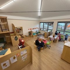 Gruppenraum mit Spiel- u. Sitzbereichen Das Bild zeigt einen modernen und einladenden Raum, der für Kinder gestaltet ist. An einem Tisch arbeiten mehrere Kinder an verschiedenen Aktivitäten. Die Tische sind farbenfroh und gut strukturiert. In einer Ecke befindet sich ein kleiner Spielbereich mit einer Kletterstruktur und bequemen Sitzgelegenheiten. Große Fenster lassen viel Tageslicht herein und bieten Blick nach draußen, wo ein Spielbereich erkennbar ist. Die helle Einrichtung und die kreative Anordnung der Möbel fördern eine angenehme Lernatmosphäre.