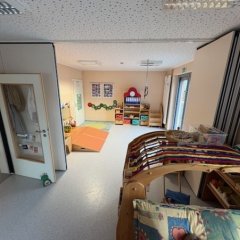 Das Bild zeigt einen Gruppenraum für Kinder. Es gibt einen hellen Boden und Wände in sanften Farben. Auf der linken Seite ist eine offene Tür zu sehen, die in einen weiteren Raum führt. Im Raum selbst gibt es Matten auf dem Boden, Spielzeug und möglicherweise Möbel wie Regale. An der Wand sind einige Bilder oder Zeichnungen befestigt. In der rechten oberen Ecke ist ein großes Fenster, das viel natürliches Licht hereinlässt, und die Raumgestaltung wirkt freundlich und einladend.