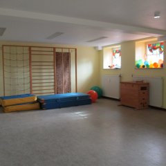 Der Raum wirkt hell und einladend mit gelben Wänden. Es gibt eine Kletterwand an einer Seite, die zum Spielen und Klettern einlädt. Auf dem Boden liegen zwei Schwammkissen in Blau und Gelb, die als weiche Unterlage dienen. In den Fenstern sind einige bunte Ballons sichtbar, die dem Raum Farbe verleihen. Zudem wird eine Holzkommode erkennbar, die wahrscheinlich für Spielmaterialien genutzt wird. Der gesamte Raum scheint für Aktivitäten und Bewegungen von Kindern gut geeignet zu sein.