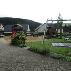 Der Spielplatz ist großzügig angelegt und bietet eine vielfältige Spielumgebung. Auf der linken Seite befindet sich eine große Rutsche und ein kleines Spielhaus in Rot, während auf der rechten Seite ein Spielgerüst mit einer Schaukel steht. Der Boden ist mit Sand und Gras bedeckt, dabei säumen Blumen und Sträucher den Platz. Im Hintergrund sind mehrere Häuser und Bäume zu sehen, die eine ruhige, ländliche Atmosphäre schaffen.