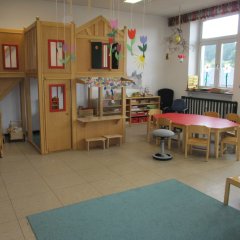Der Raum ist hell und freundlich gestaltet, mit einem großen Holzspielhaus in der Ecke, das über eine Treppe erreichbar ist. Es gibt eine kleine Küche und Fenster mit bunten Vorhängen. In der Mitte steht ein runder Tisch mit Stühlen, die zum Spielen und Arbeiten einladen. Um den Tisch herum sind farbenfrohe Spielgeräte sichtbar, und es hängt eine Deko aus Blumen und anderen Motiven von der Decke. Der Boden ist mit einem blauen Teppich bedeckt, der eine gemütliche Atmosphäre schafft. Die gesamte Einrichtung ist kinderfreundlich und fördert kreatives Spielen.