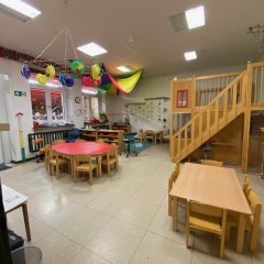Der Raum auf dem Bild ist ein Kindergarten-Spielraum. Er ist hell erleuchtet und großzügig gestaltet. In der Mitte stehen verschiedene Tische und Stühle, darunter ein großer runder Tisch. In einer Ecke gibt es eine Kletter- oder Spielstruktur, die den Kindern zusätzliche Möglichkeiten zum Spielen und Entdecken bietet. Die Wände sind farbenfroh dekoriert, und es gibt eine Vielzahl von Spielmaterialien, darunter bunte Teller, die von der Decke hängen. Der gesamte Raum strahlt eine einladende und freundliche Atmosphäre aus, die zum Spielen und Lernen anregt.