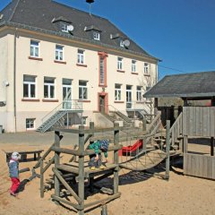 Das Bild zeigt einen Spielplatz vor dem großen, mehrstöckigen KiTa-Gebäude. Auf dem Spielplatz befindet sich eine Holz-Kletterstruktur mit Rutschen und Seilen, die den Kindern verschiedene Spielmöglichkeiten bietet. Zwei Kinder spielen aktiv, eines klettert und das andere befindet sich in der Nähe der Struktur. Der Boden ist mit Sand bedeckt, und der klare blaue Himmel deutet auf einen sonnigen Tag hin, was eine fröhliche und einladende Atmosphäre für Aktivitäten im Freien schafft.