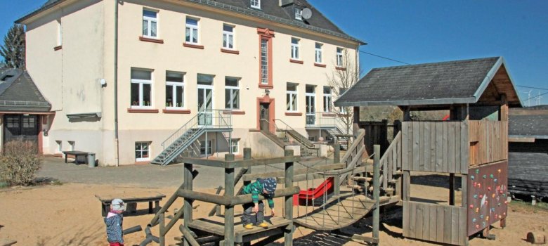 Das Bild zeigt einen Spielplatz vor dem großen, mehrstöckigen KiTa-Gebäude. Auf dem Spielplatz befindet sich eine Holz-Kletterstruktur mit Rutschen und Seilen, die den Kindern verschiedene Spielmöglichkeiten bietet. Zwei Kinder spielen aktiv, eines klettert und das andere befindet sich in der Nähe der Struktur. Der Boden ist mit Sand bedeckt, und der klare blaue Himmel deutet auf einen sonnigen Tag hin, was eine fröhliche und einladende Atmosphäre für Aktivitäten im Freien schafft.