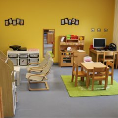 Der Raum auf dem Bild ist ein Spiel- oder Gruppenraum für Kinder. Die Wände sind in einem warmen Gelb gestrichen, was eine freundliche Atmosphäre schafft. In der Mitte stehen ein Tisch mit Stühlen und ein großer grüner Teppich. Es gibt verschiedene Spielmöglichkeiten, darunter eine Küche mit Spielutensilien und ein Computer, der wohl für die kindliche Nutzung gedacht ist. Weiche Stühle und Regale mit Spielzeug und Materialien runden die Einrichtung ab, die zum Spielen und Lernen anregt. Die große Fensterfront lässt viel Tageslicht herein, was den Raum zusätzlich heller und einladender macht.