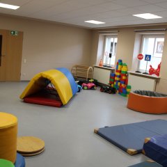 Der Raum auf dem Bild ist ein Bewegungs- und Spielraum für Kinder. Er ist hell und großzügig gestaltet, mit mehreren Fenstern, die viel Tageslicht hereinlassen. In dem Raum befinden sich verschiedene Kletter- und Turnelemente, wie ein aufblasbares Tunnelzelt und große, bunte Schaumstoffelemente zum Spielen und Entdecken. Außerdem liegen einige Krabbelfahrzeuge und Bauklötze auf dem Boden, die zur motorischen Förderung und zum kreativen Spielen einladen. Die farbenfrohe Ausstattung sorgt für eine anregende und spielerische Atmosphäre.
