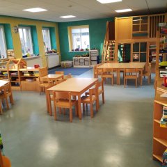 Der Raum zeigt einen freundlichen Gruppenraum. Es gibt mehrere Holztische und Stühle, die für Gruppenaktivitäten oder Einzelarbeiten genutzt werden können. Regale mit Büchern und Spielzeug sind vorhanden, was auf eine spielerische Lernumgebung hindeutet. Die Wände sind in hellen Farben gestrichen, wodurch eine fröhliche Atmosphäre entsteht. Im Hintergrund ist eine kleine Treppe sichtbar, möglicherweise zu einer Spielebene oder Ausruhecke. Insgesamt wirkt der Raum hell, gut organisiert und fördert die Kreativität der Kinder.