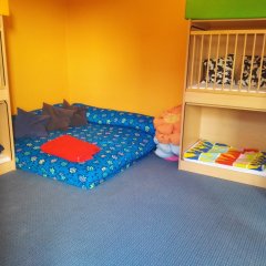 Der Raum ist eine gemütliche Ruhe- oder Schlafzone für Kinder. Es gibt zwei Etagenbetten mit farblich passenden Kissen, die für eine einladende Atmosphäre sorgen. Die Wände sind in einem hellen Gelb gestrichen, was den Raum freundlich wirken lässt. Auf dem Boden liegt ein blauer Teppich, der zusätzlichen Komfort bietet. In einer Ecke befindet sich eine große Liegefläche mit bunten Kissen und Decken, die zum Relaxen einlädt. Insgesamt strahlt der Raum eine ruhige und entspannende Umgebung aus.