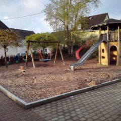 Der Spielplatz bietet eine vielfältige Outdoor-Spielumgebung für Kinder. In der Mitte steht ein Spielturm mit einer Rutsche und einem Kletterbereich. Auf der linken Seite befindet sich eine Schaukel. Der Boden ist mit Holzspänen bedeckt, was eine sichere Landefläche bietet. Außerdem sind die umgebenden Wände aus Holz, die eine geschützte Atmosphäre schaffen. Grüne Bäume und Büsche sorgen für einen naturbelassenen Look und Schatten, wodurch der Spielplatz einladend und abenteuerlich wirkt.
