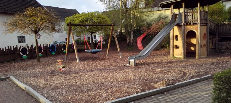 Der Spielplatz bietet eine vielfältige Outdoor-Spielumgebung für Kinder. In der Mitte steht ein Spielturm mit einer Rutsche und einem Kletterbereich. Auf der linken Seite befindet sich eine Schaukel. Der Boden ist mit Holzspänen bedeckt, was eine sichere Landefläche bietet. Außerdem sind die umgebenden Wände aus Holz, die eine geschützte Atmosphäre schaffen. Grüne Bäume und Büsche sorgen für einen naturbelassenen Look und Schatten, wodurch der Spielplatz einladend und abenteuerlich wirkt.