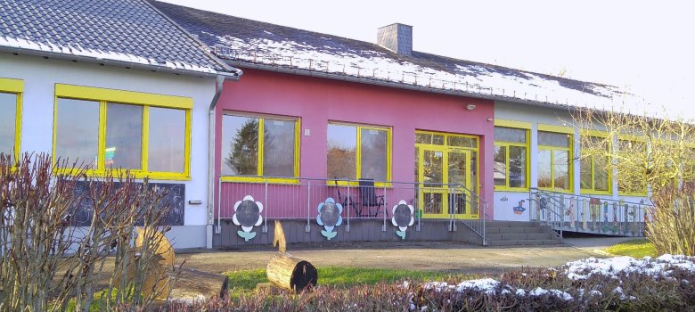 Das Gebäude der Kindertagesstätte Weinsheim ist eine einzelgeschossige Einrichtung. Die Fassade ist in hellen Farben gestaltet, mit einem pinkfarbenen Teil und weißen Wänden, die von leuchtend gelben Fensterrahmen umgeben sind. Große Fenster sorgen für ausreichend Licht und einen einladenden Blick nach außen. Vor dem Gebäude sind Blumenillustrationen und gestaltete Geländer sichtbar, die die kinderfreundliche Atmosphäre unterstreichen.