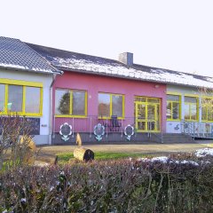 Das Gebäude der Kindertagesstätte Weinsheim ist eine einzelgeschossige Einrichtung. Die Fassade ist in hellen Farben gestaltet, mit einem pinkfarbenen Teil und weißen Wänden, die von leuchtend gelben Fensterrahmen umgeben sind. Große Fenster sorgen für ausreichend Licht und einen einladenden Blick nach außen. Vor dem Gebäude sind Blumenillustrationen und gestaltete Geländer sichtbar, die die kinderfreundliche Atmosphäre unterstreichen.