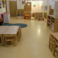 Der  Gruppenraum für Kinder ist hell und freundlich. Es gibt mehrere Tische mit kleinen Stühlen, die geeignet für Kinder sind.  Auf der linken Seite befindet sich eine Spielstation mit Spielzeugkisten und einem Spielhaus. Die Wände sind mit kindgerechten Kunstwerken oder Bildern dekoriert. Der Boden ist hell und hat einfache, leicht zu reinigende Oberflächen. Insgesamt wirkt der Raum einladend und fördert kreative Aktivitäten.