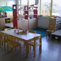 Der Raum ist freundlich und gut beleuchtet, ideal für Kinderaktivitäten. Es gibt einen zentralen Tisch mit kleinen Stühlen, die zum Spielen oder Arbeiten geeignet sind. Offene Regale an den Wänden präsentieren Spielzeug und Materialien für verschiedene Aktivitäten. Ein Bereich mit farbigen Regalen und einem hochgelegenen Podest bietet Raum zum Spielen und Entdecken.  Große Fenster lassen viel natürliches Licht herein und bieten Blick nach außen, was den Raum hell und einladend macht. Insgesamt vermittelt der Raum eine positive und förderliche Umgebung für Kinder.