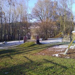Der Spielplatz ist weitläufig und naturbelassen, ideal für Kinder zum Spielen und Entdecken. Es gibt eine Rutsche, die sanft ins Rasenland führt, sowie eine Wippe, die das Spiel bereichert. Ein Klettergerüst aus Holz steht zur Verfügung, das den Kindern Bewegung und Herausforderungen bietet. Der Boden ist größtenteils mit Rasen und Holzspänen bedeckt, was die Sicherheit beim Spielen erhöht. Umgeben von Bäumen bietet der Platz sowohl Schatten als auch eine naturnahe Atmosphäre. Der Spielplatz schafft eine einladende Umgebung, die aktives Spielen und soziale Interaktionen fördert.