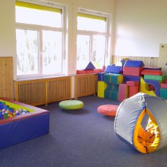 Der Tobraum ist ein hell und freundlich gestalteter Spielbereich. In der linken Ecke befindet sich ein aufgefüllter Pool mit bunten Bällen. An der Wand stehen bunte, weiche Bausteine in verschiedenen Formen, die Kinder zum Spielen und Bauen einladen. Auf dem Boden liegen verschiedene runde Polster, die als Sitzgelegenheit dienen. An einer Wand ist eine Kletterfläche mit bunten Griffen angebracht, die für Bewegung und Abenteuer sorgt. Große Fenster lassen viel Tageslicht herein, was den Raum freundlich wirken lässt. Insgesamt ist die Atmosphäre lebhaft und einladend, ideal für spielerisches Lernen und Bewegungsfreiheit.