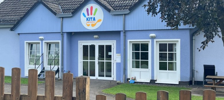 Das Bild zeigt das Eingangsgebäude der Kita Winterspelt. Die Fassade ist in einem freundlichen blauen Farbton gehalten, und über der Eingangstür befindet sich ein buntes Schild mit dem Schriftzug "KITA". Vor dem Gebäude steht ein Holzlattenzaun, der den Eingang umgibt. Die Fenster des Hauses sind groß, was einen hellen und einladenden Eindruck vermittelt. Im Hintergrund sind einige Grünflächen und Bäume zu sehen, die eine angenehme Umgebung schaffen.     