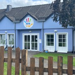 Das Bild zeigt das Eingangsgebäude der Kita Winterspelt. Die Fassade ist in einem freundlichen blauen Farbton gehalten, und über der Eingangstür befindet sich ein buntes Schild mit dem Schriftzug "KITA". Vor dem Gebäude steht ein Holzlattenzaun, der den Eingang umgibt. Die Fenster des Hauses sind groß, was einen hellen und einladenden Eindruck vermittelt. Im Hintergrund sind einige Grünflächen und Bäume zu sehen, die eine angenehme Umgebung schaffen.     