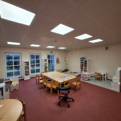 Gruppenraum mit Leseecke, Spielzeugen und Büchern Das Bild zeigt das Innere eines Gruppenraums in einer Kindertagesstätte. Der Raum ist hell und freundlich, mit großen Fenstern, die Tageslicht hereinlassen. Auf dem Boden liegt ein roter Belag, und der Raum ist mit einzelnen Tischen und Stühlen ausgestattet. Es gibt eine Leseecke mit einem Teppich und Regalen, die mit Spielzeugen und Büchern gefüllt sind. Die Decke hat moderne, flache Leuchten, die für eine angenehme Beleuchtung sorgen. Der Raum wirkt einladend und kindgerecht gestaltet.