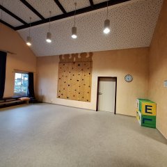 Das Bild zeigt einen großen, freundlichen Gruppenraum. Die Wände sind in einem warmen Beigeton gestrichen. An einer Wand befindet sich eine Kletterwand mit bunten Griffen, die zu Bewegung und Spiel einlädt. Der Raum hat hohe Decken mit modernen Pendelleuchten. Eine Uhr und ein bunter Kasten mit Buchstaben stehen ebenfalls im Raum. Der Boden ist mit einem hellen Linoleumbelag versehen, und es gibt eine gemütliche Sitzgelegenheit an einem Fenster. Der Raum bietet Platz für verschiedene Aktivitäten und Spiele.