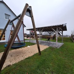 Das Bild zeigt einen Außenbereich einer Kindertagesstätte mit einem Spielplatz. Im Vordergrund befindet sich eine Großschaukel mit einer runden Netzschaukel. Die Spielfläche ist mit Sand und kleinen Kieselsteinen gestaltet, die eine sichere Unterlage bieten. Im Hintergrund sieht man ein Pavillon, der Schatten und Schutz bietet. Das Gebäude der Kita ist nur teilweise sichtbar und wird von einer grünen Wiese umgeben. Der Bereich wirkt einladend und kindgerecht, ideal für Aktivitäten im Freien.